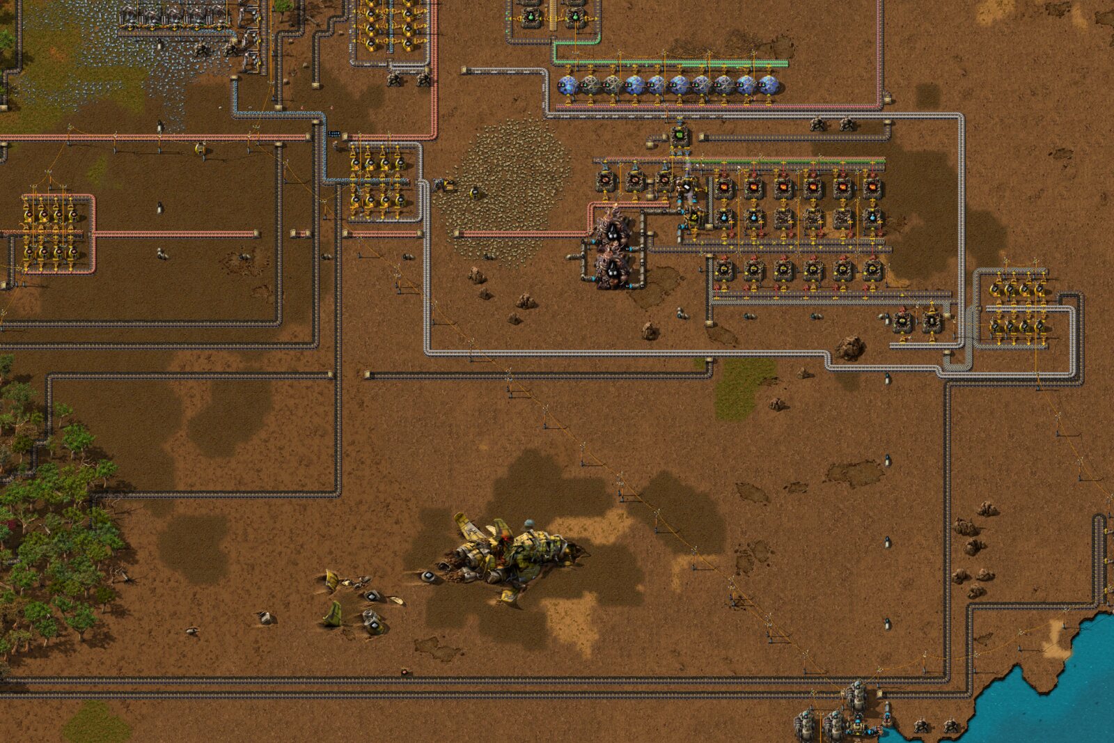 Overhead view of a Factorio base on brown ground: smelting arrays and grey belts on the left, rows of assembling machines at center right with a row of labs above them, a crashed spaceship wreck near the bottom, and a lake at the lower right.